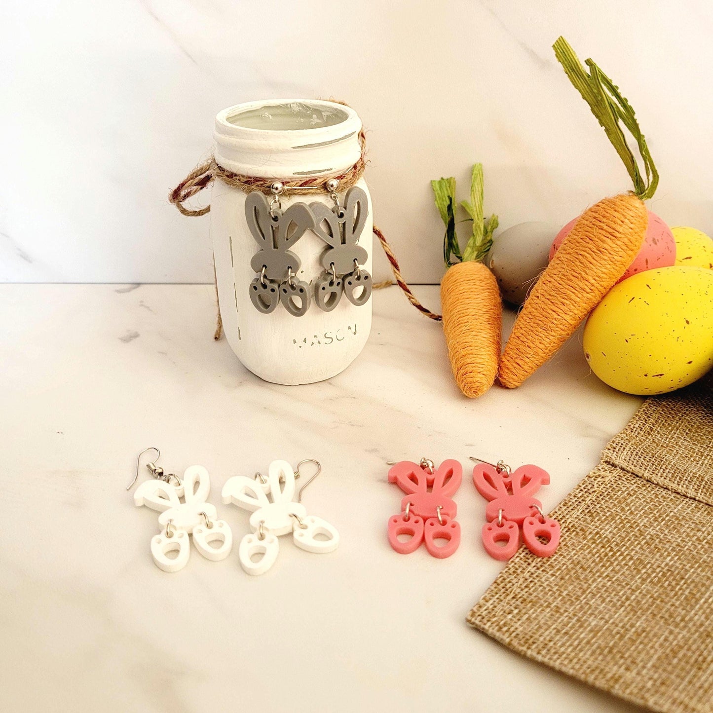 3 pairs of earrings in grey, pink and white acrylic. The grey pair is hanging from a small painted white jar. The earrings are bunny ears with 2 feet that dangle from the bottom.
